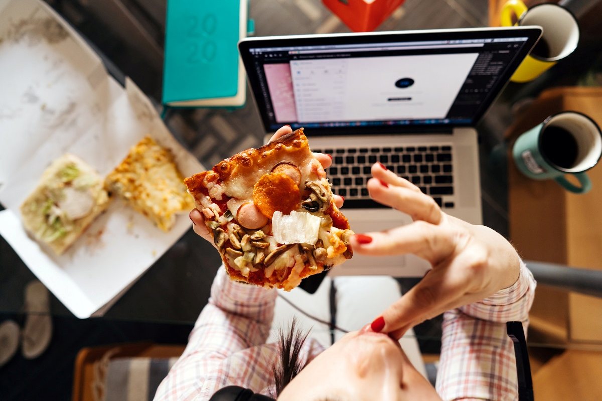 mãos segurando pedaço de pizza e computador a frente