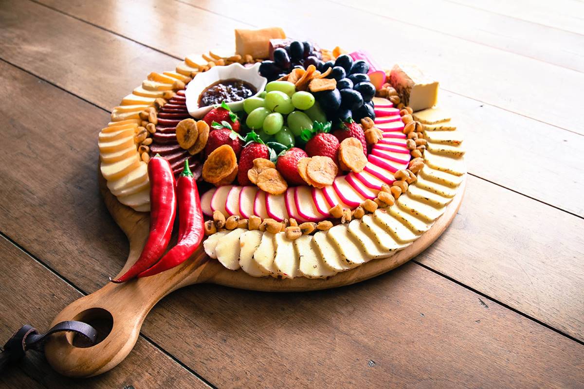 Imagem colorida de uma tábua de frios, contendo queijos, frutas e embutidos