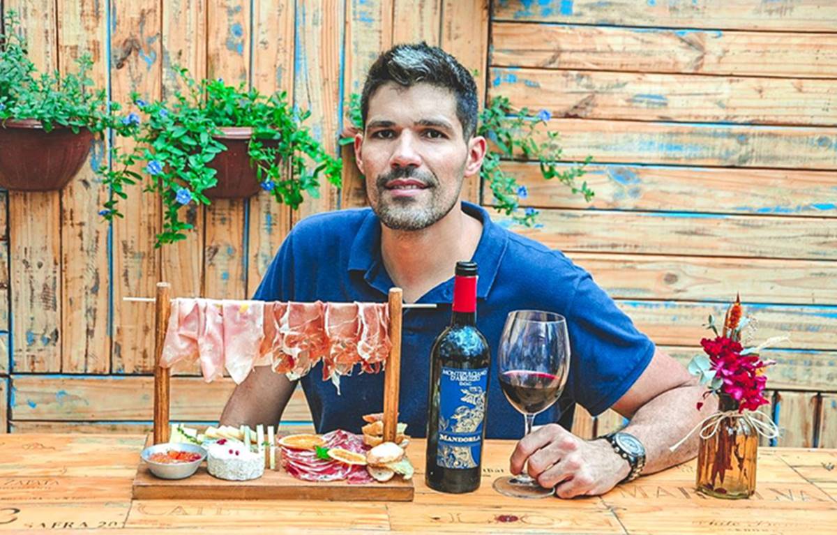 Homem segurando uma taça de vinho que está sobre uma mesa de madeira junto com uma garrafa de vinho e uma tábua de frios