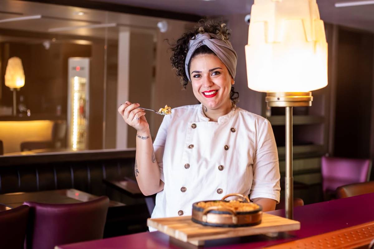 Mulher com cabelos cacheados vestida com uma dolmã de chef e segurando um garfo com comida