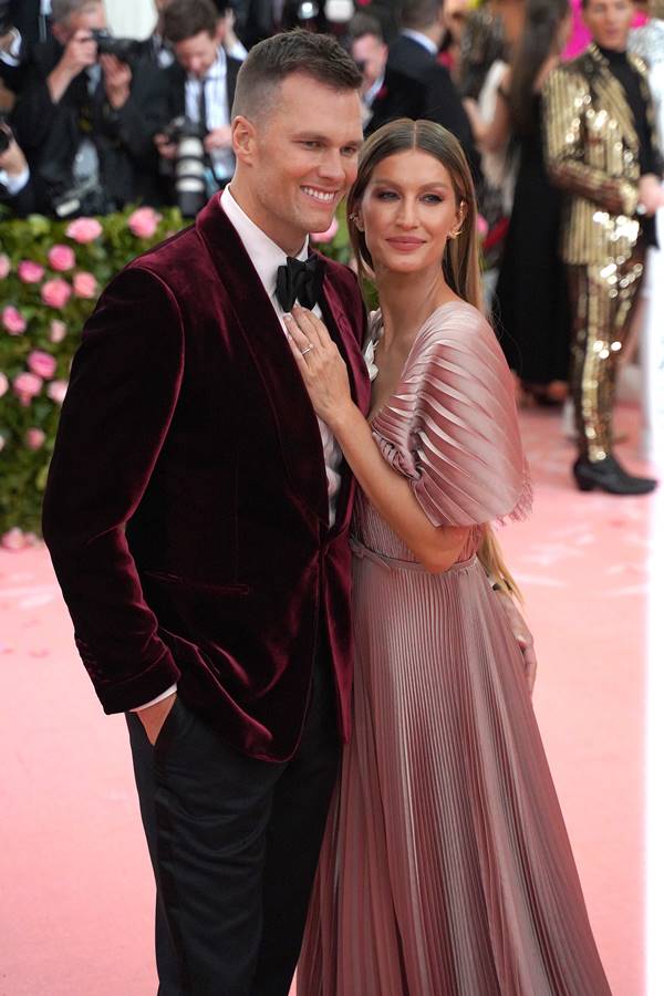 Foto colorida de Tom Brady e Gisele Bündchen. Eles estão sorridentes e abraçados. Ele usa um terno todo na cor vinho e ela um vestido esvoaçante rosa - Metrópoles