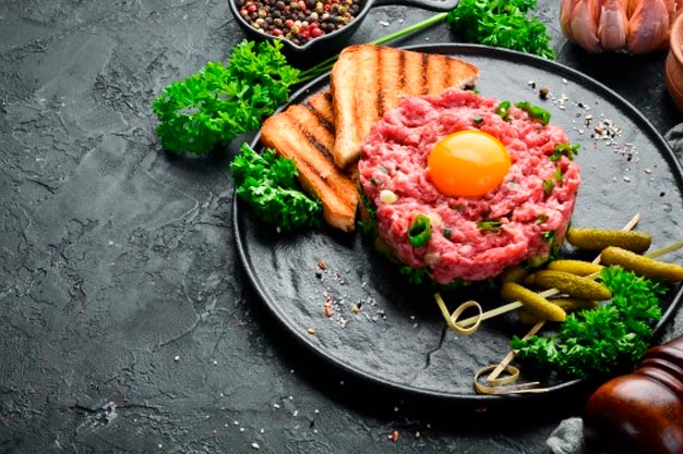 A foto mostra um prato cinza com Steak Tartare, receita que leva carne crua e gema de ovo; Ao lado, torradinhas e folhagens verdes comestíveis