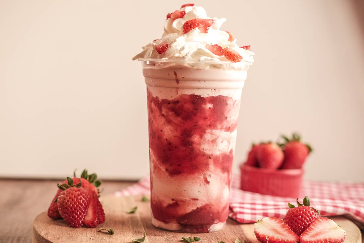 Na fotografia vemos uma pequena tabua com um copo de plastico cheio de milkshake de morango, que criam camadas, e por cima chantilly com pedaços de morango. Ao lado, morangos e no fundo uma toalha adrez branco e vermelha sob uma pequena vasilha com morangos