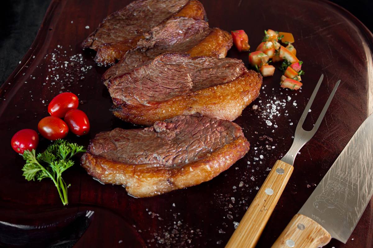 Fotografia de tábua de madeira com fatias de carne assada com bordas de gordura. Com sal salpicado dos lados, um garfo de churrasco e um faca no canto inferior direito e tomatinhos e folhinhas de salsa no canto inferior esquerdo