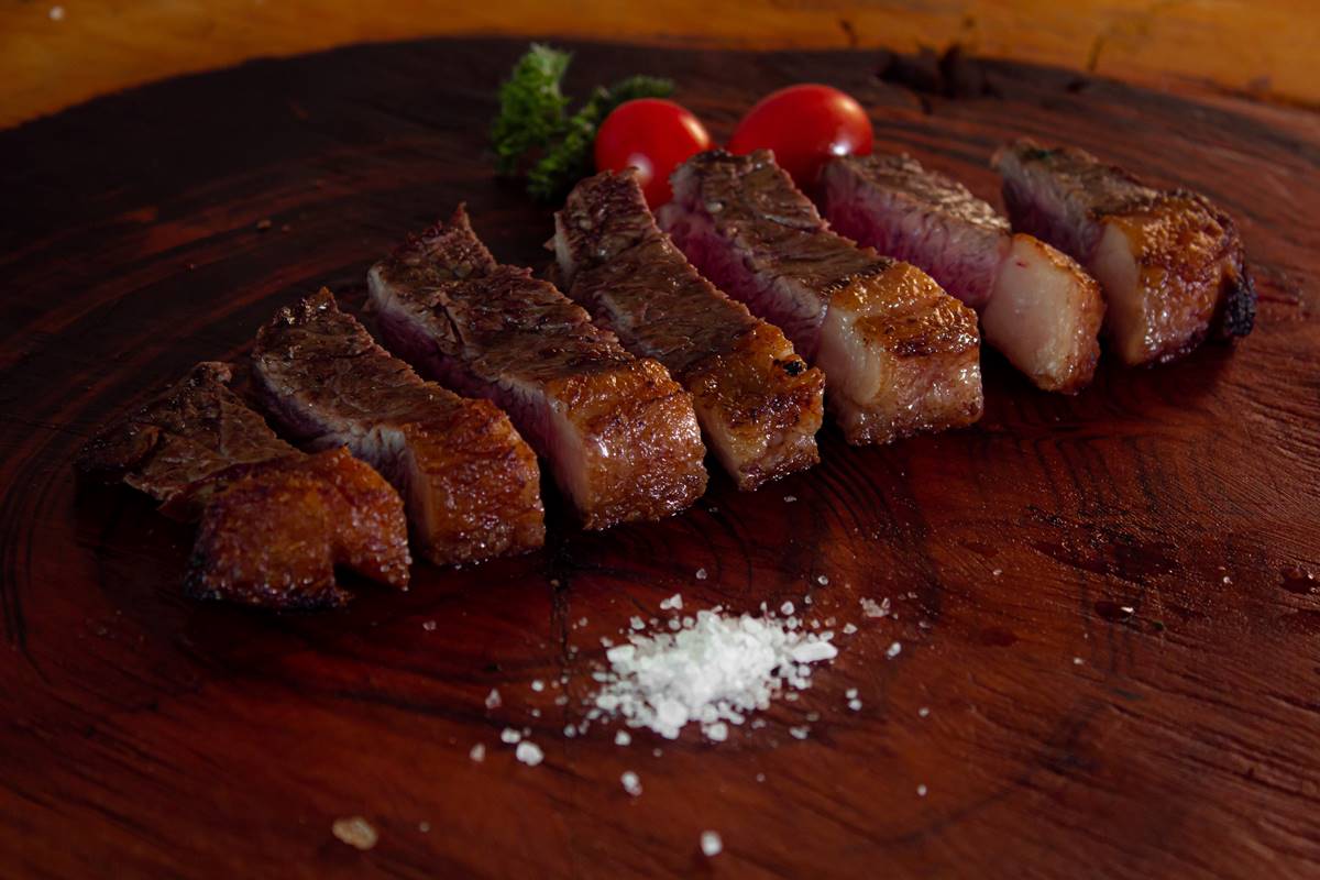 Fotografia de tábua de madeira com carne cortada em tirinhas, com um montinho de sal no centro inferior e tomatinhos e folhas de salsa no centro superior, atrás da carne fatiada