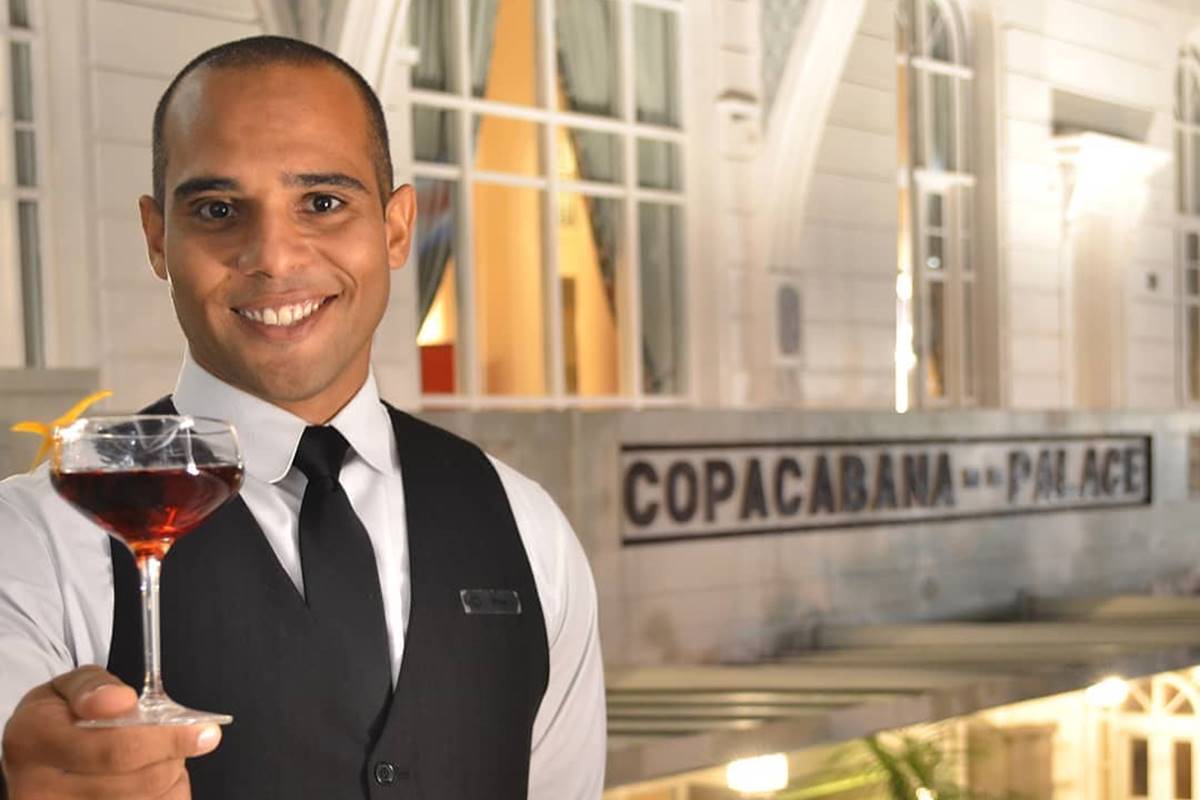 Na foto vemos a facha do hotel cobacana palace em cinza e no canto esquerdo um homem de camisa branca, colete e gravata pretos segurando uma taça com drinque marrom