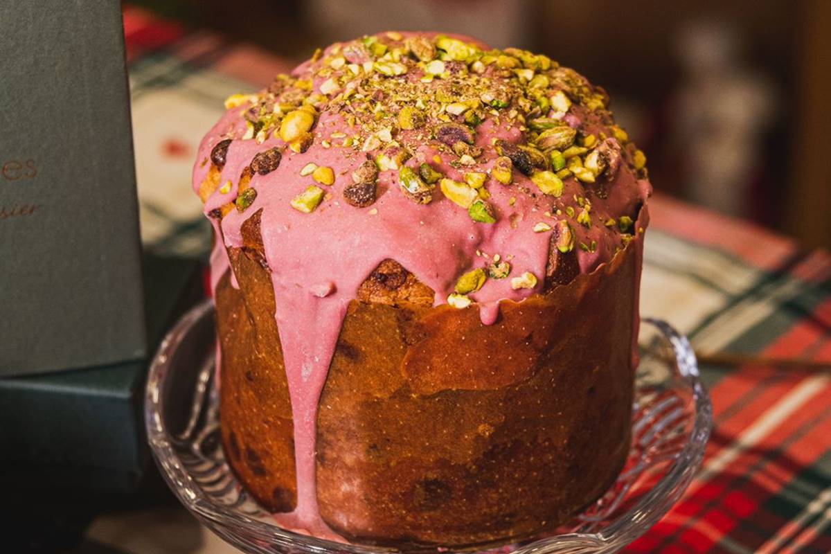 Na foto temos um chocotone sobre uma bailarina de vidro com cobertura de chocolate rosa e pistache