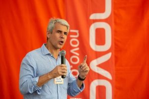 Fotografia colorida de Felipe D'ávila. Ele é um homem branco, de cabelos lisos e grisalhos penteados para o lado. Na foto, aparece falando diante de um microfone, com o dedo indicador da mão direita levantado. Veste camisa azul-claro. O fundo desfocado é alanjado e lê-se Novo