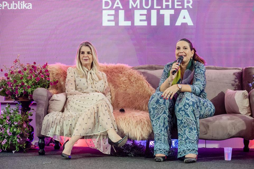 Duas mulheres sentadas, uma com roupa azul e segura um microfone