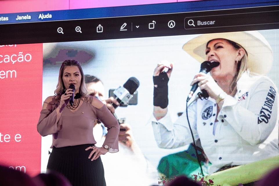 Mulher fala ao microfone. Ao fundo há um telão com uma foto de uma cowgirl 