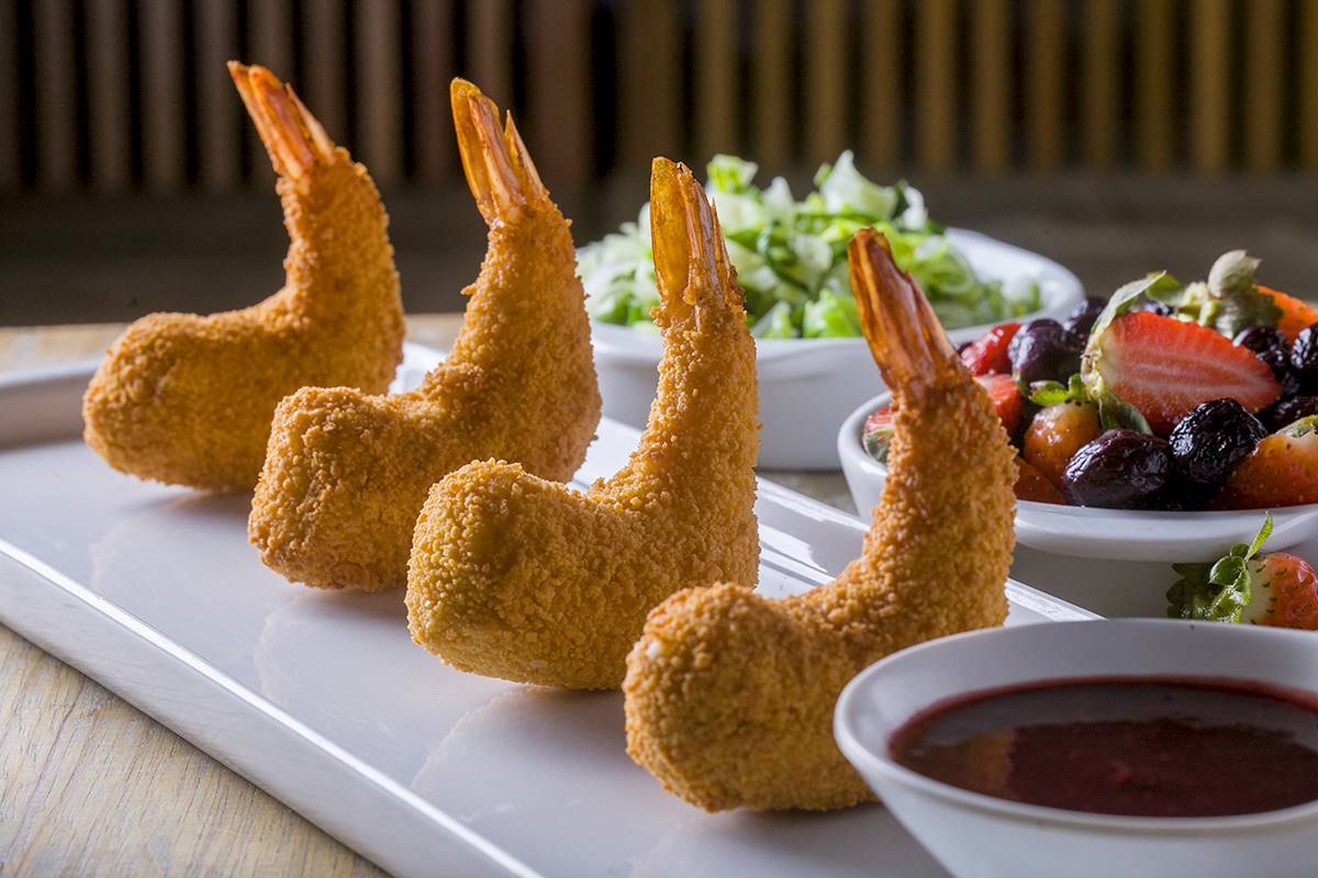 Na foto temos um prato retangular com camaroes empanados e um potinho de molho e ao fundo duas cumbucas brancas, uma com salada e outra com frutas