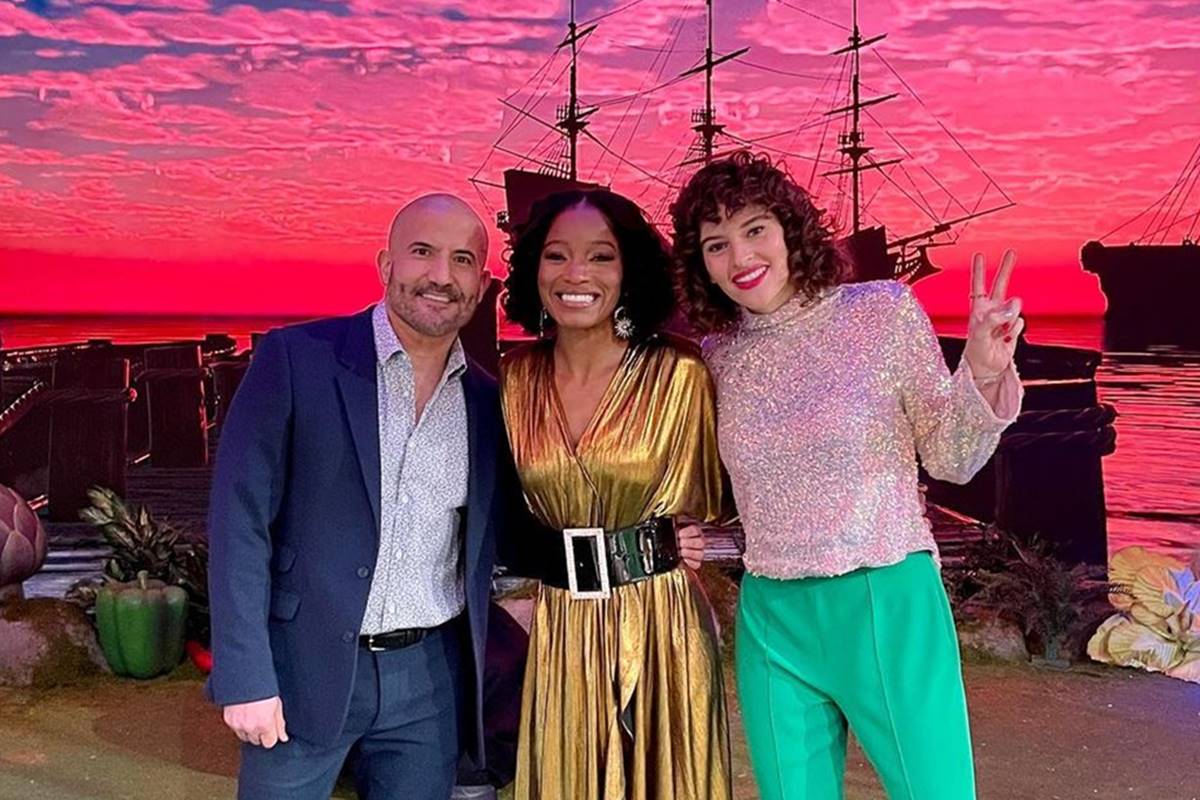 No fundo da foto temos um céu rosa e um barco pirata e na frente um homem careca de terno azul, uma mulher negra de vestido dourado e uma mulher branca com blusa brilhosa e calça verde