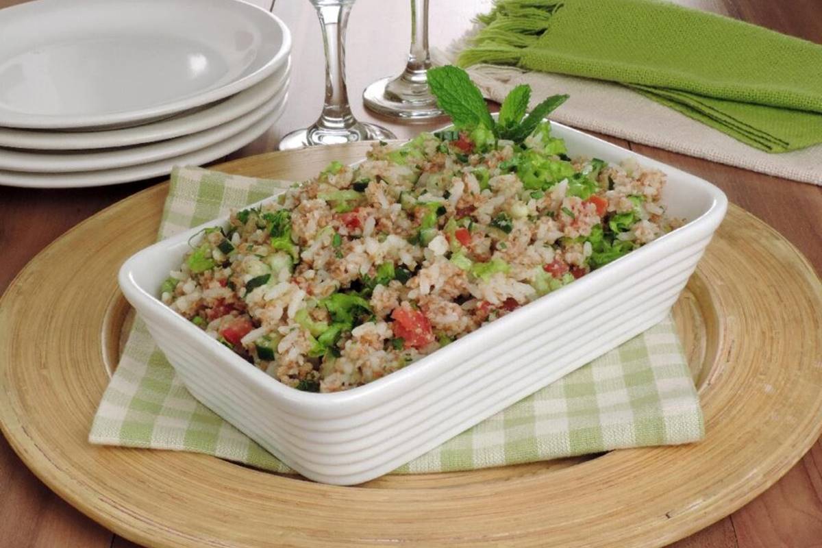 Na foto temos um suplar amadeirado com uma toalha xadrez verde e branca e uma travessa branca de ceramica com tabule de arroz dentro