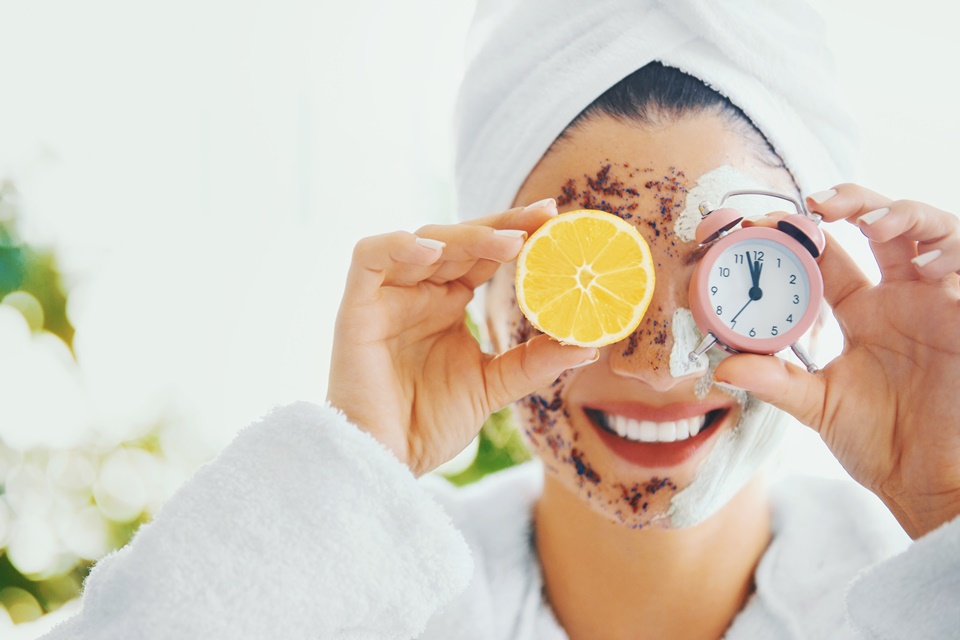 Mulher com toalha na cabeça e produto de beleza no rostosegura relógio e metade de laranja em frente aos olhos