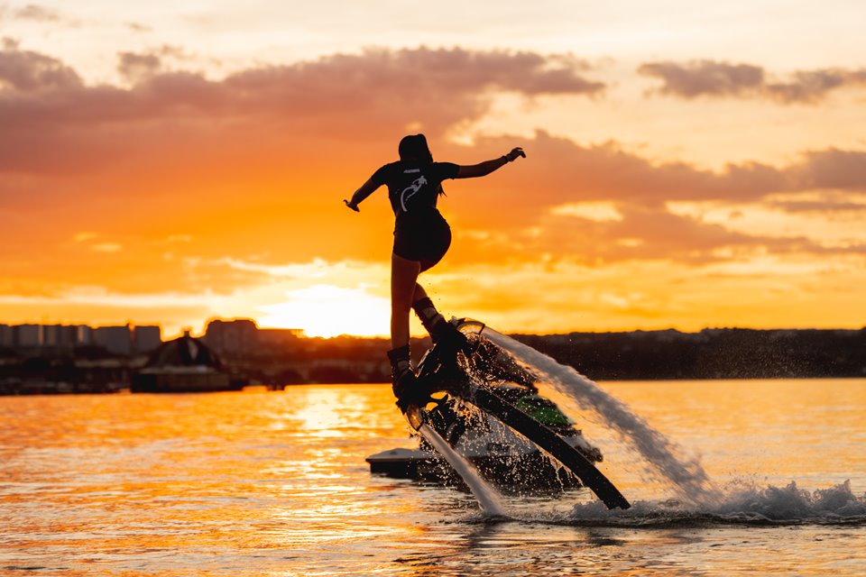 Mulher usando objeto que a faz voar sob as águas com pôr do sol ao fundo