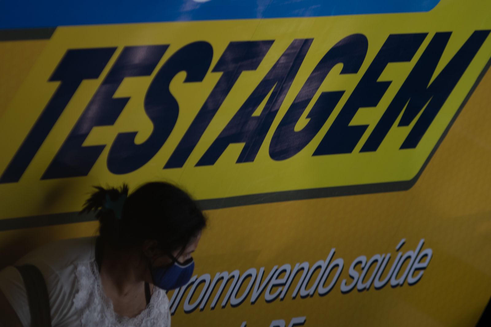 Mulher em frente a uma placa gigante amarelo escrito "testagem"