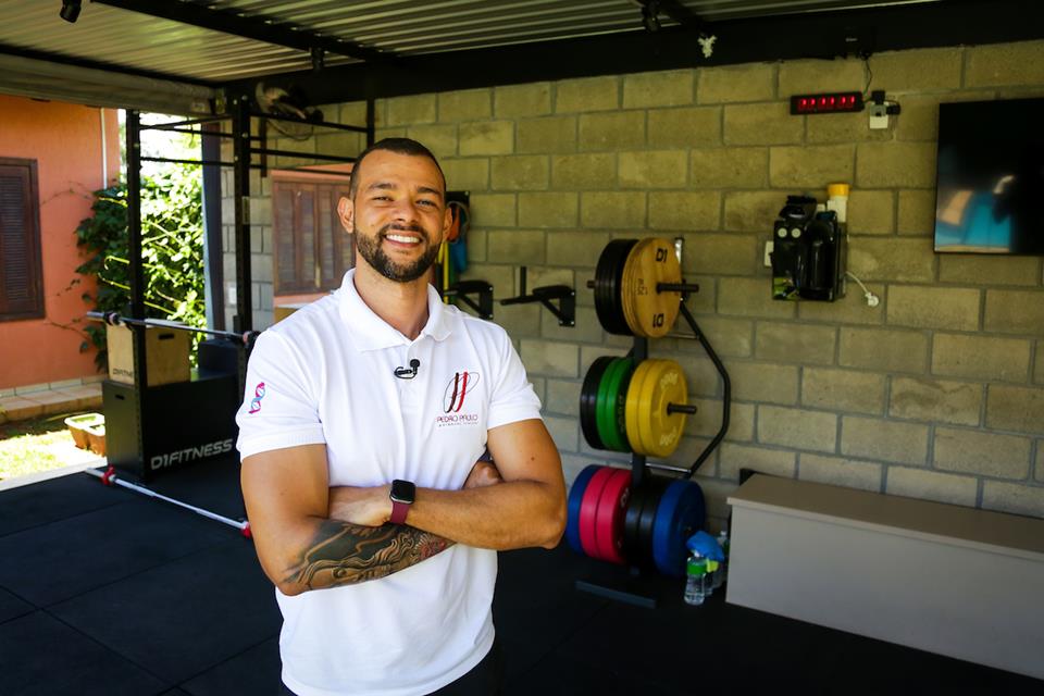 Homem de braços cruzados. Ao fundo da foto, equipamentos de academia