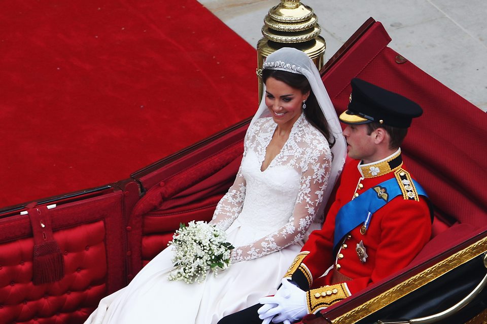 Mulher vestida de noiva e com buquê. O homem está com trajes militares. Os dois estão em uma charrete de luxo