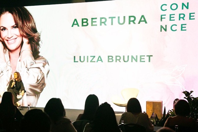 Painel com o nome Luiza Brunet escrito. Mulher sentada à esquerda