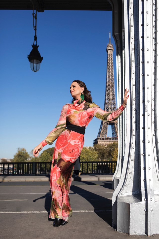 Torre Eiffel ao fundo de imagem. Mulher está escorada em uma pilastra. Ela está com vestido em tons de verde, rosa e vermelho