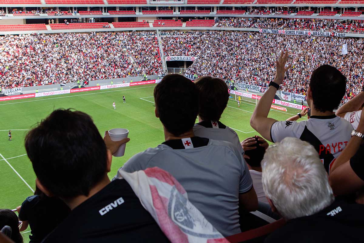 Foto do estádio com torcedores de costas vibrando