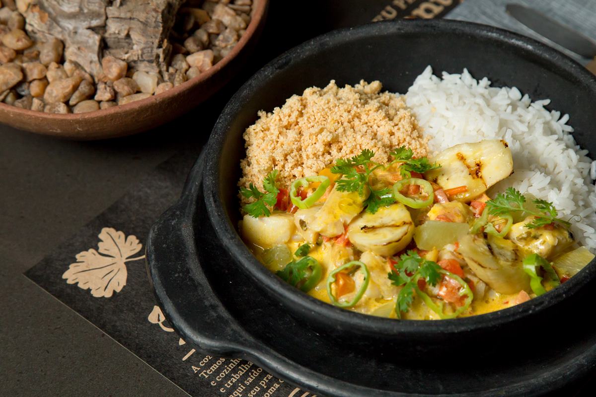 Na foto temos uma travessa de barro preta com arroz, farofa e moqueca de peixe 