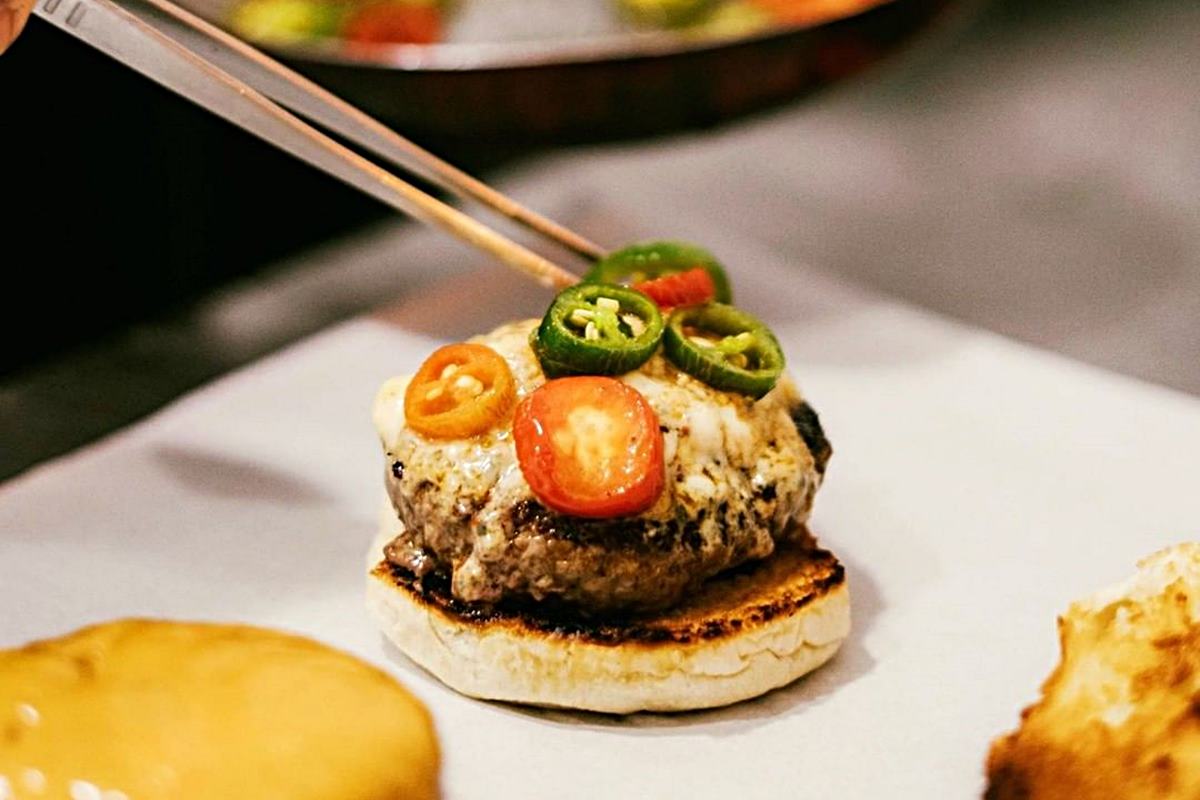 Na foto temos um hamburguer e uma pinça colocando rodelas de pimentas