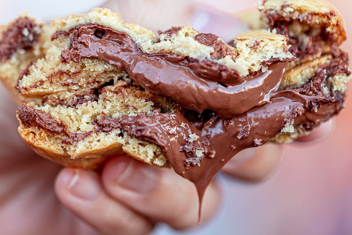 Na foto temos um cookie partido ao meio com uma metade por cima da outra e recheio de creme de nutella escorrendo