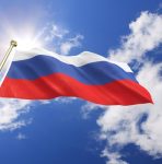 A imagem mostra a bandeira da Rússia sob ceu com nuvens e um Sol brilhando atrás dessa - Metrópoles