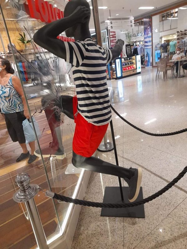 Manequim preto com blusa listrada e bermuda vermelha quebrando a vitrine de vidro da marca Reserva, em corredor de shopping