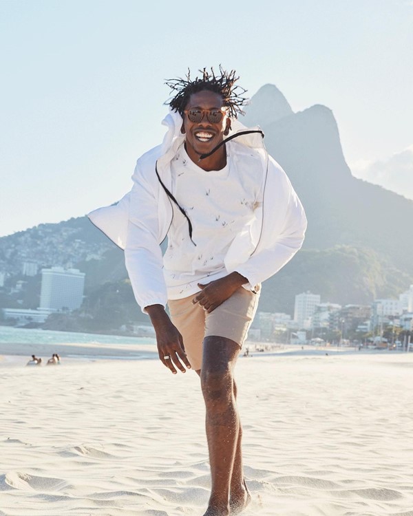 Homem negro com casaco e blusa branca e short bege. Ele está pulando na praia, no Rio de Janeiro, com o pão de açúcar ao fundo