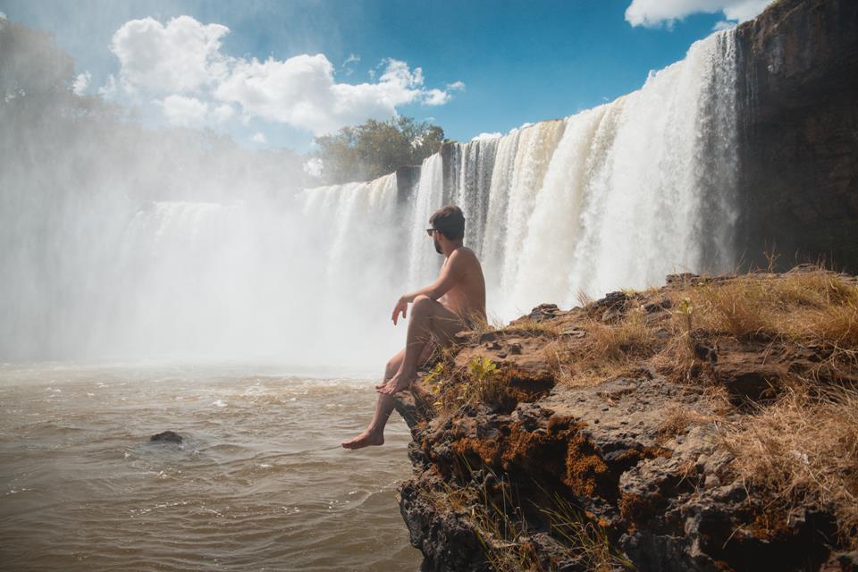 Homem sentado em uma pedra. Ao fundo, há uma cachoeira