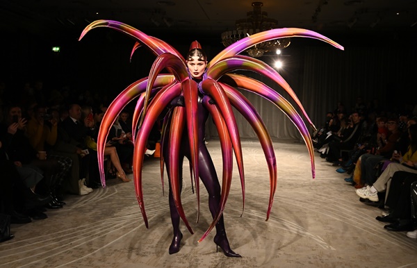 Modelo branca com cabelo preto na passarela. Ela veste uma estrutura inflável que parece uma planta, mas é vermelha e roxa.