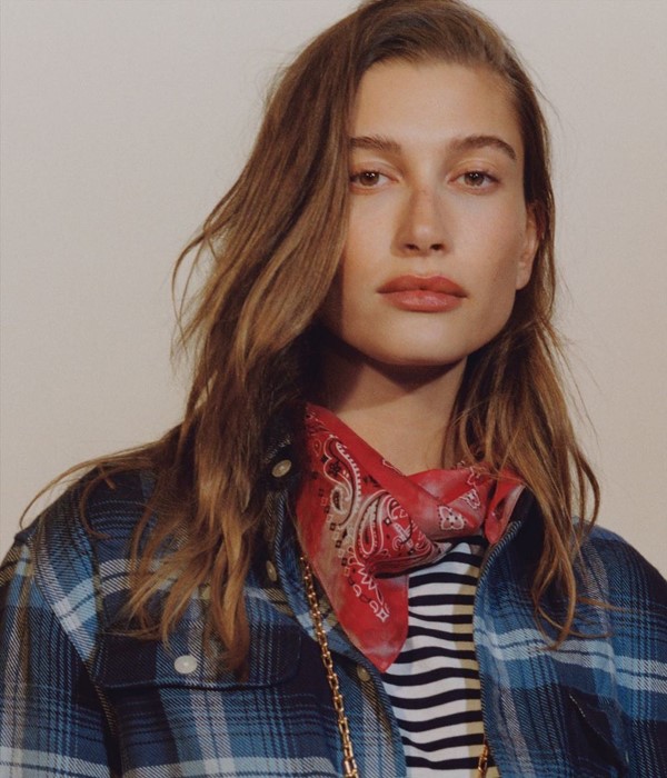 Mulher branca e loura com blusa listrada, camisa xadrez e uma bandana vermelha no pescoço. Ela é a modelo Hailey Bieber, casada com o cantor Hailey Bieber