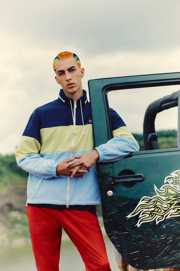 Homem branco modelo posando com roupas da marca Lacoste. Ele possui o cabelo tingido e está posando ao lado de um carro.