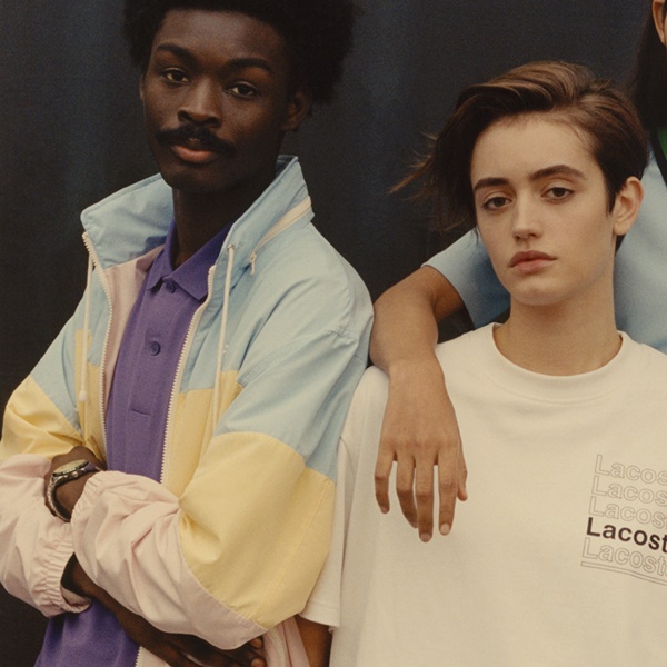 Modelos posam usando roupas da Lacoste. O primeiro modelo é um homem negro e veste uma jaqueta listrada com cores pastel. A segunda é uma modelo branca, de cabelo curto, com uma camiseta branca.