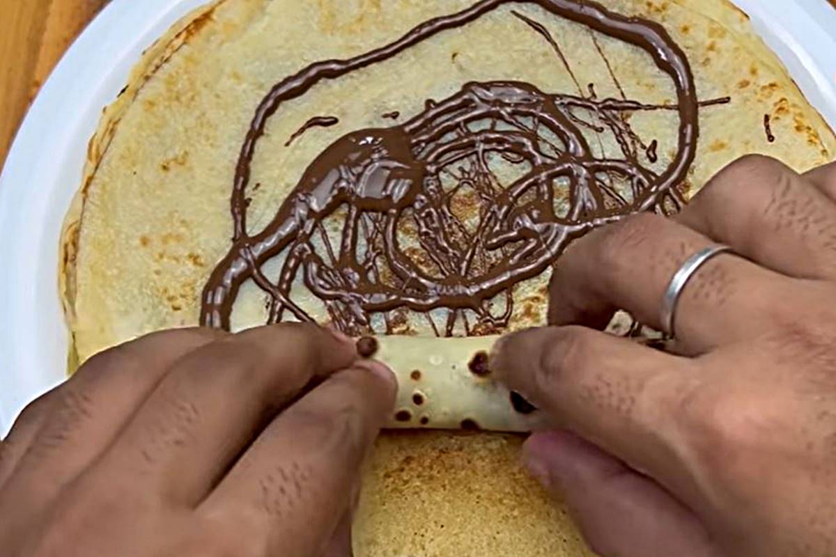 na foto vemos duas mãos enrolando uma panqueca coberta de chocolate