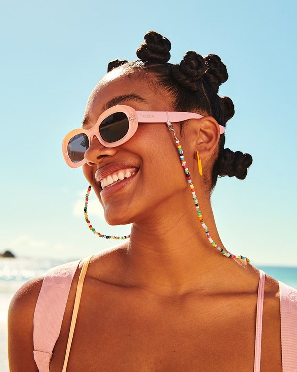 Mulher negra sorrindo com óculos escuros na cor rosa. Ao fundo, é possível ver o mar