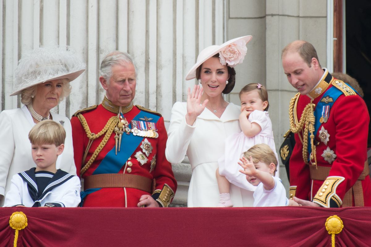 Foto colorida. Camilla Parker, príncipe Charles, Kate Middleton, princesa Charlotte, príncipe George e príncipe William. Eles estão em uma varanda. Os homens vestem trajes militares