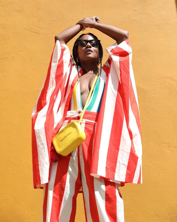 Mulher negra com roupa calça e kimono de listras brancas e vermelhas. Ela também usa uma bolsa quadrada amarela da marca Balenciaga. Ela posa em parede amarela.