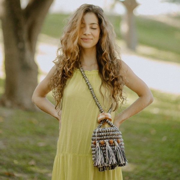 Mulher branca com cabelos cacheados claros. Ela usa um vestido amarelo e uma bolsa transversal em crochê da marca Catarina Mina. Ao fundo é possível ver um jardim