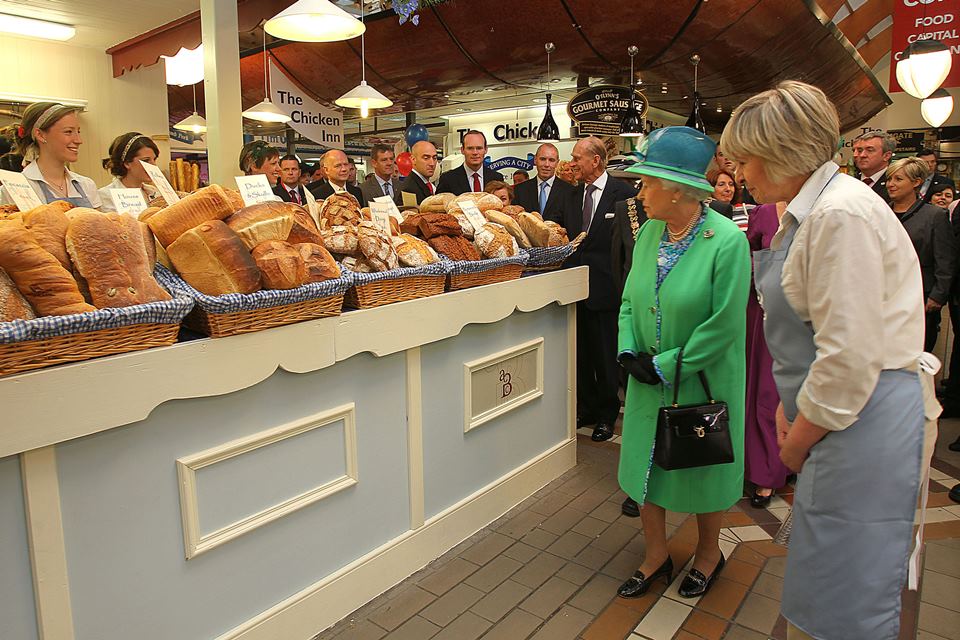 Foto colorida. Rainha Elizabeth com vestido verde. Ela olha pães