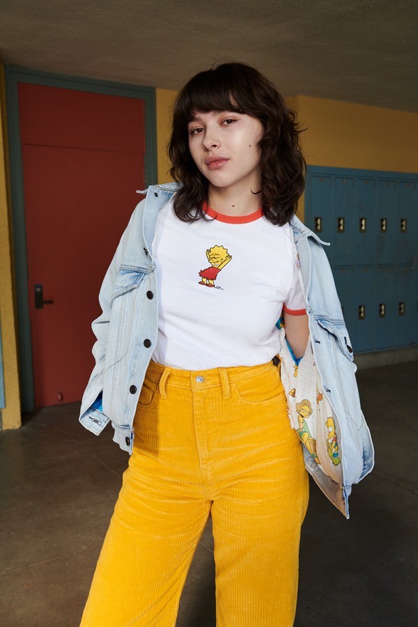 Mulher branca com cabelo preto curto de franja. Ela está de blusa branca, calça amarela e jaqueta jeans. As peças são da marca Levi's em parceria com o desenho Os Simpsons.