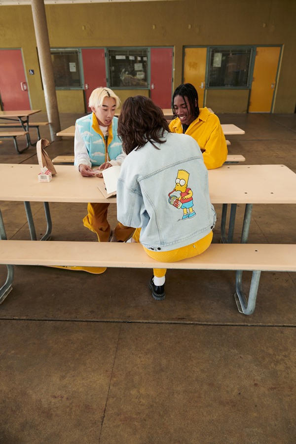 Mulher branca sentada em mesa com homem negro e outro asiático. Eles estão sentados em uma mesa do refeitório da escola fazendo refeição. Todos usam peças da marca Levi's em parceria com o desenho Os Simpsons.