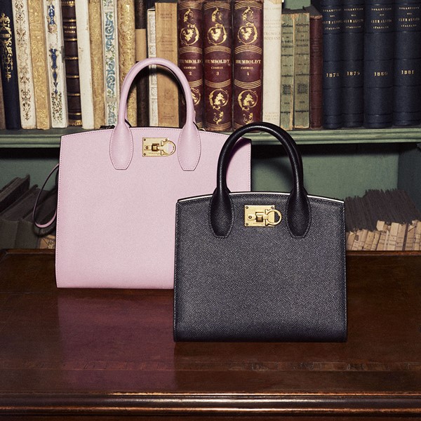 Bolsas rosa e preta da marca Salvatore Ferragamo. Elas estão apoiadas em uma mesa de madeira escura e, ao fundo, é possível ver prateleiras com livros.