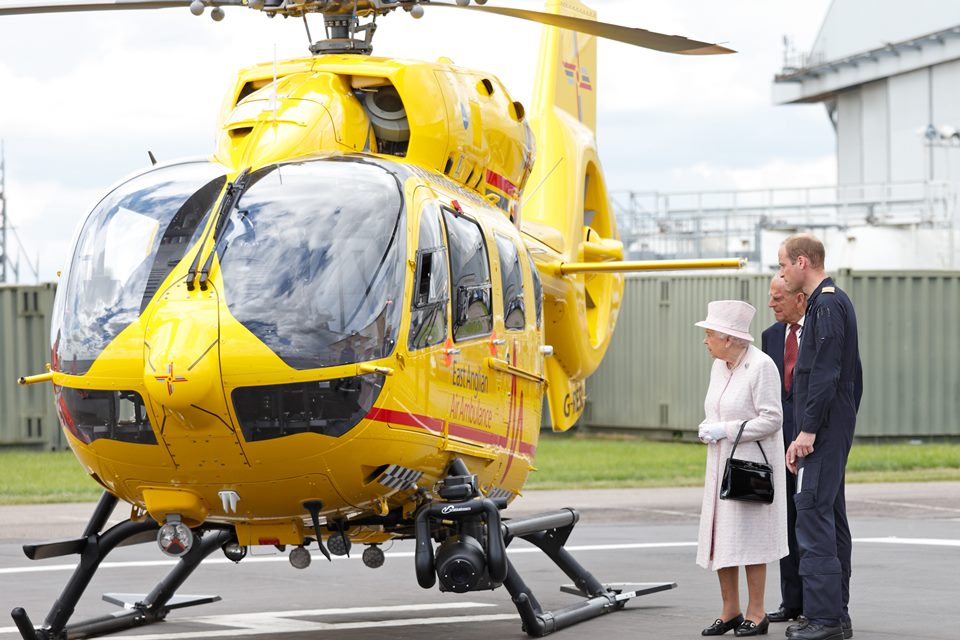 Foto colorida. Rainha Elizabeth, príncipe William e príncipe Philip perto de um helicóptero amarelo