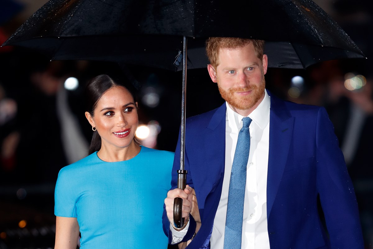 Foto colorida. Meghan Markle e príncipe Harry seguram um guarda-chuva. Meghan usa um vestido vermelho e Harry, terno