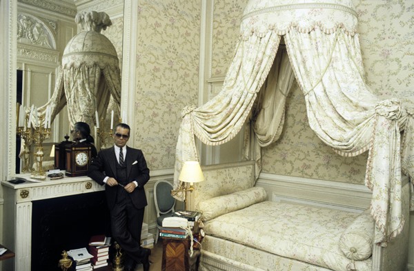 Estilista Karl Laferfeld jovem posando em quarto decorado. Ele usa um terno preto e óculos escuros, mesmo dentro do cômodo.