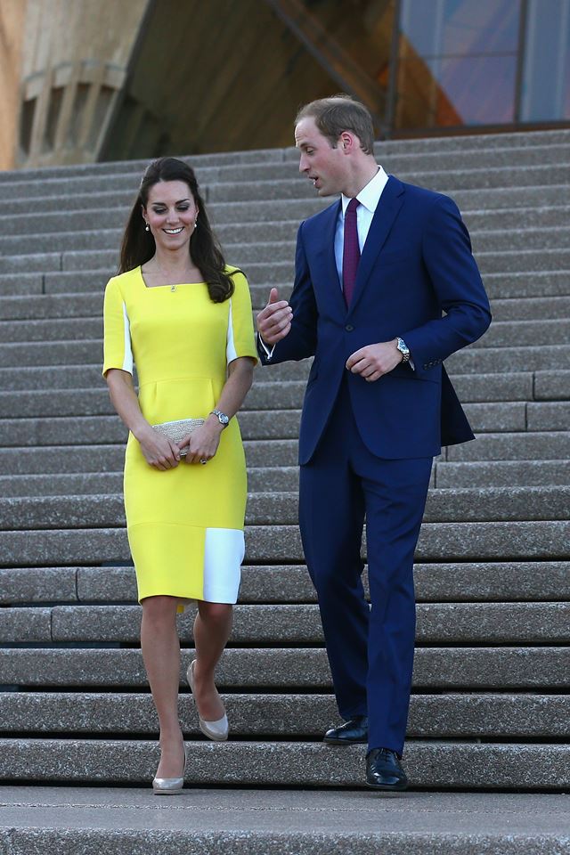 Foto colorida. Kate Middleton e príncipe William descem uma escadaria. Ela usa um vestido amarelo e ele, um terno