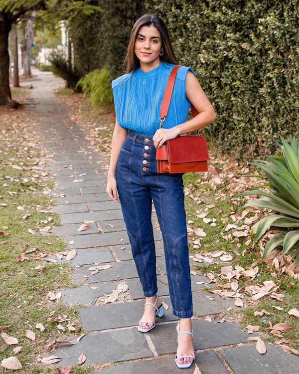 Mulher branca com cabelo castanho liso e cumprido usando calça jeans e blusa azul. Ela carrega uma bolsa marrom e calça um salto alto azul claro. Atrás é possível ver um jardim.